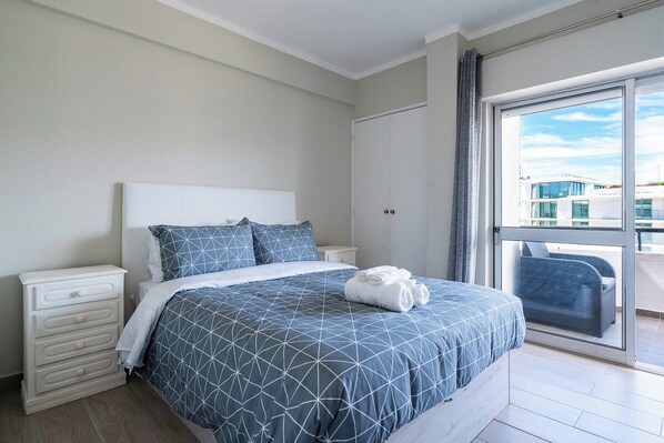 A bedroom with a bed, a nightstand, a dresser, and a view through a large window.