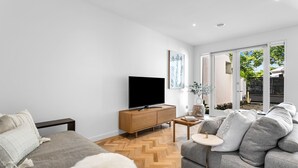 A modern living room with a sofa, a TV, a wooden cabinet, and a herringbone floor. - Bev's Retreat by Ready Set Stay - peaceful retreat and close to all attractions. (South Yarra)