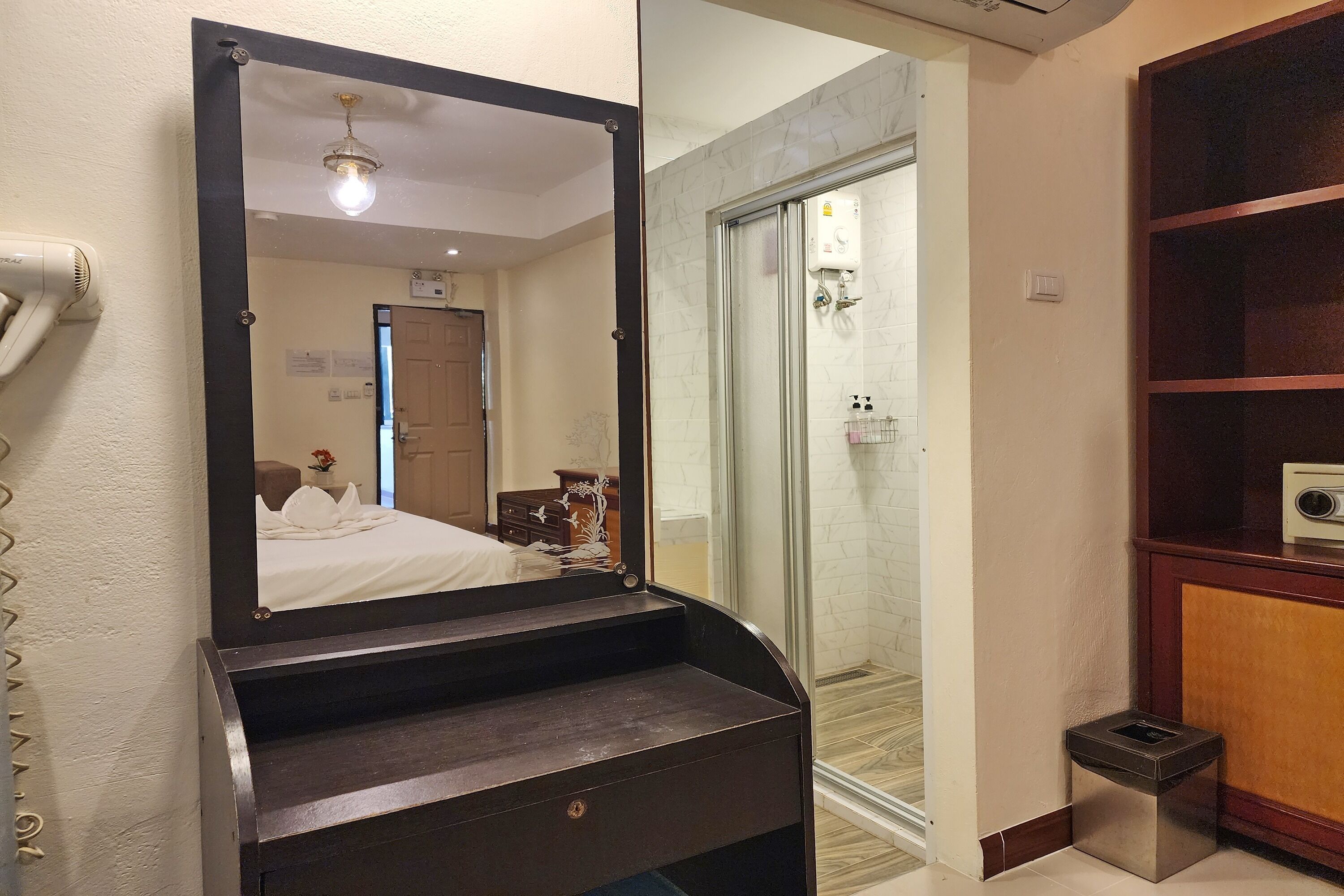 A hotel bathroom with a large mirror, a hairdryer, a shower, and a small trash bin.
