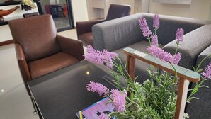 Ein modernes Wohnzimmer mit einem Glastisch, einem braunen Ledersessel, einem grauen Sofa und einem Holztisch mit lila Blumen.