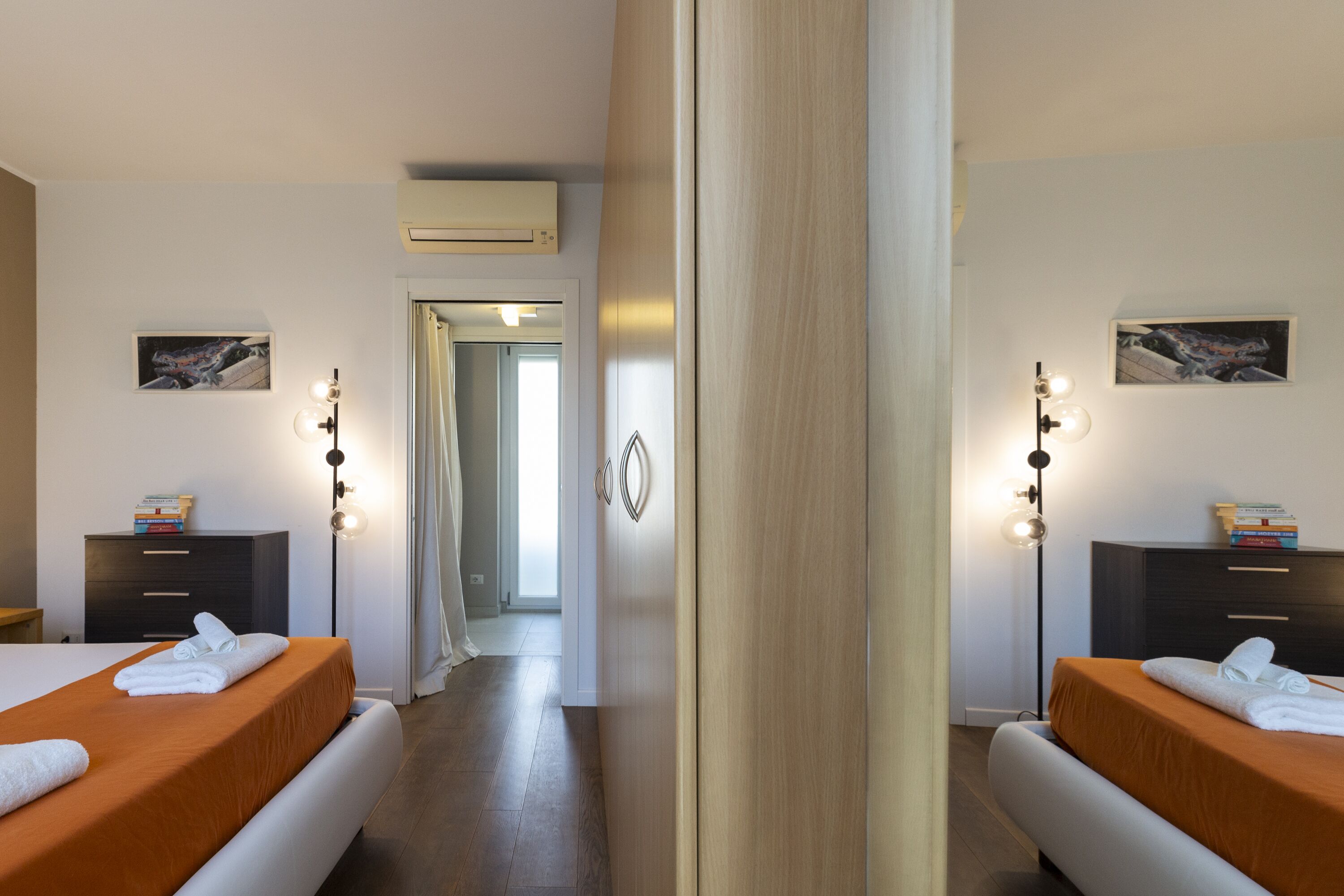 A hotel room with two beds, a wardrobe, a desk, and a mirror. There is a door leading to another room and an air conditioning unit on the wall.