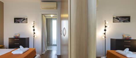 A hotel room with two beds, a wardrobe, a desk, and a mirror. There is a door leading to another room and an air conditioning unit on the wall.
