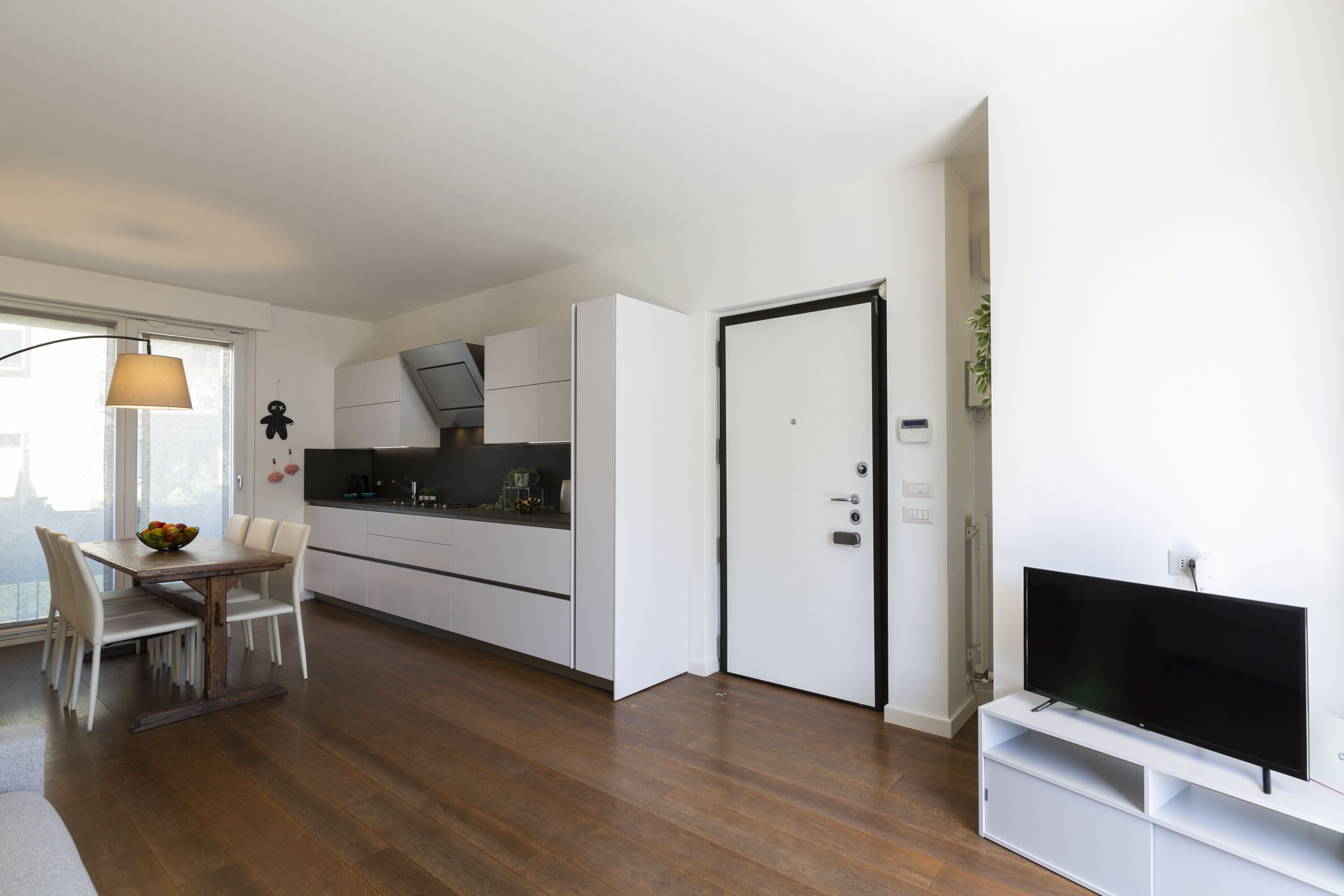 A modern kitchen with white cabinets, a dining table with chairs, and a flat-screen TV mounted on a white stand.