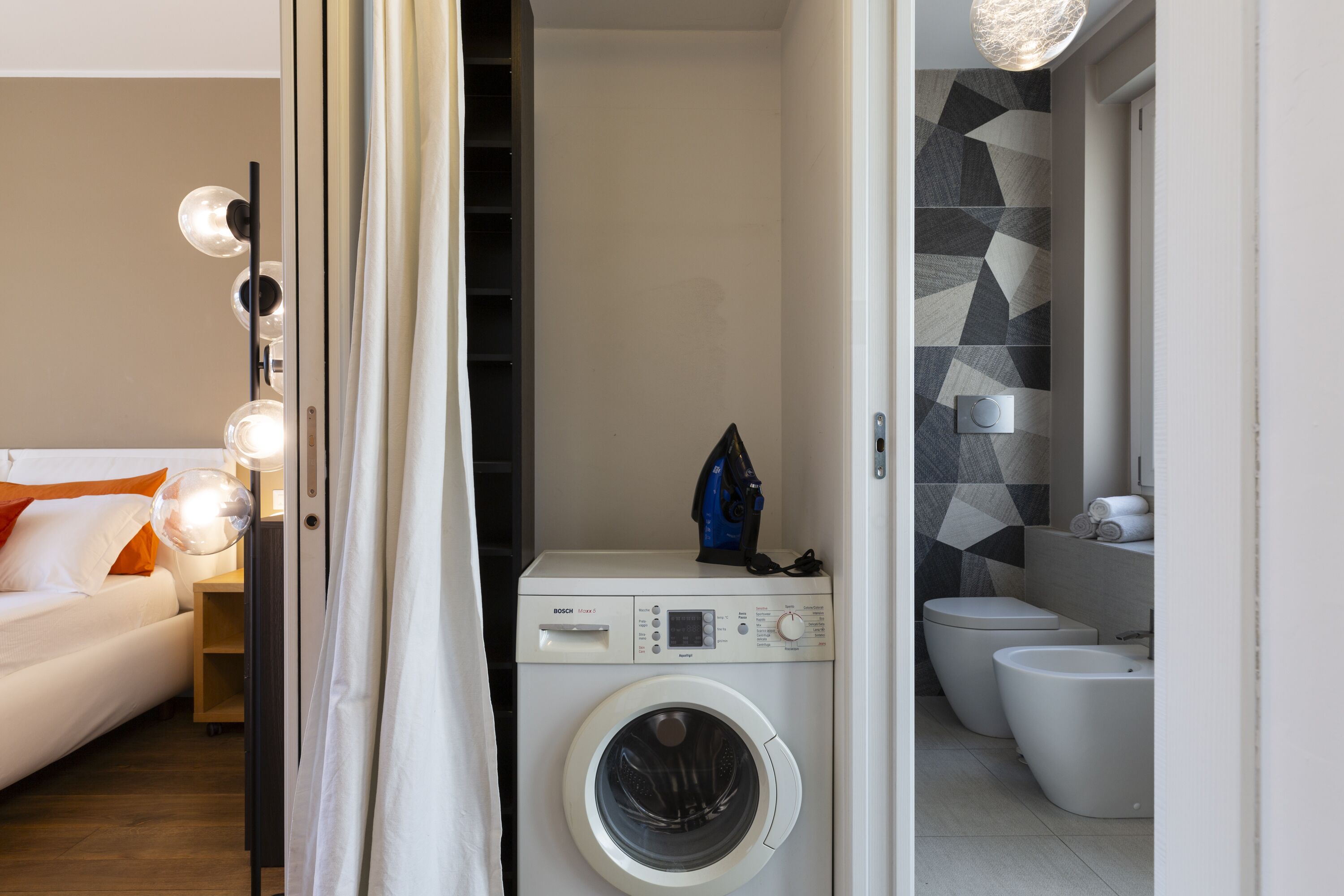 Een moderne hotelkamer met een wasmachine, een bed met oranje kussens, een nachtkastje met een lamp en een badkamer met toilet en bidet.