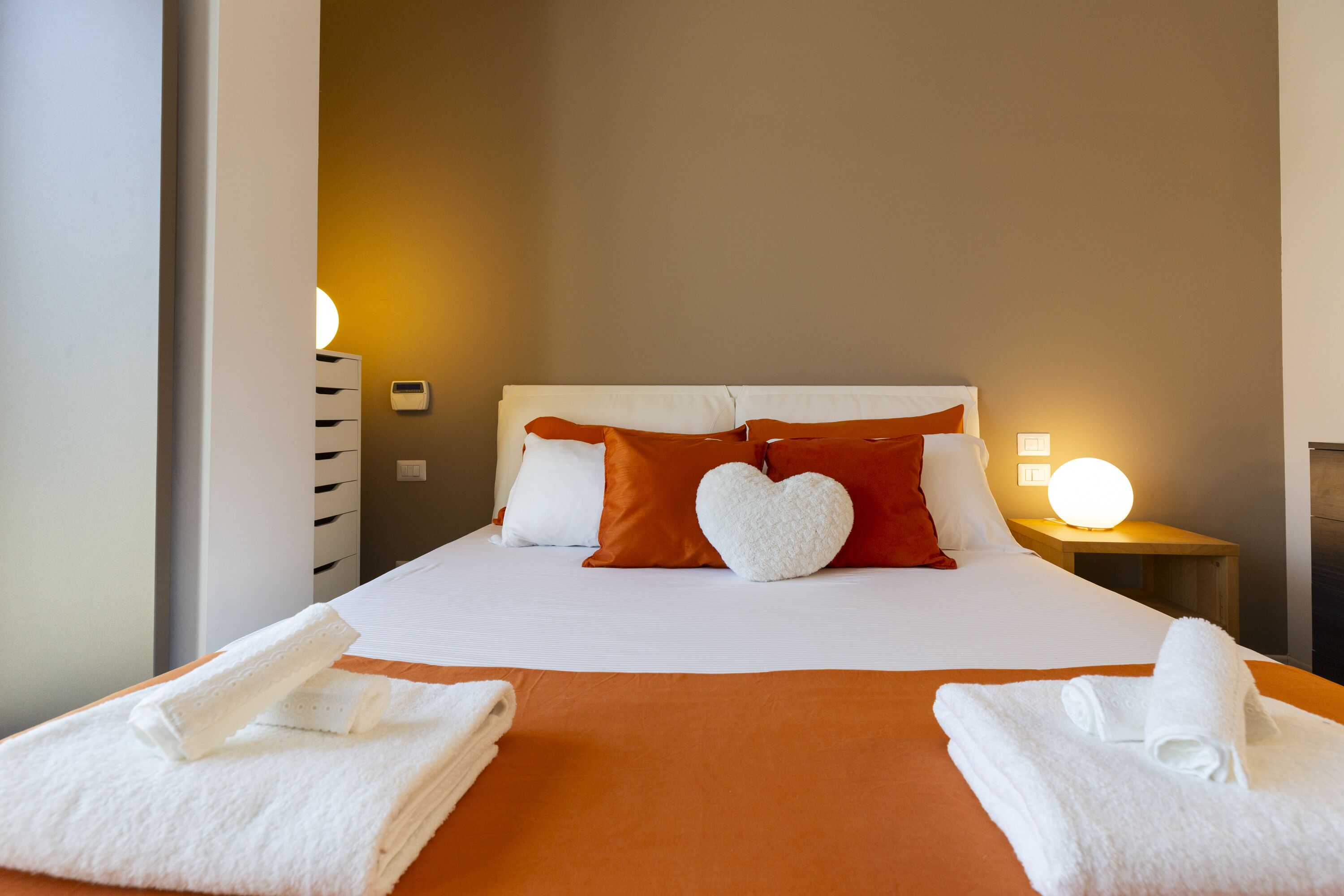 Una cama bien hecha con ropa de cama blanca y naranja, una almohada con forma de corazón y toallas dobladas a un lado.