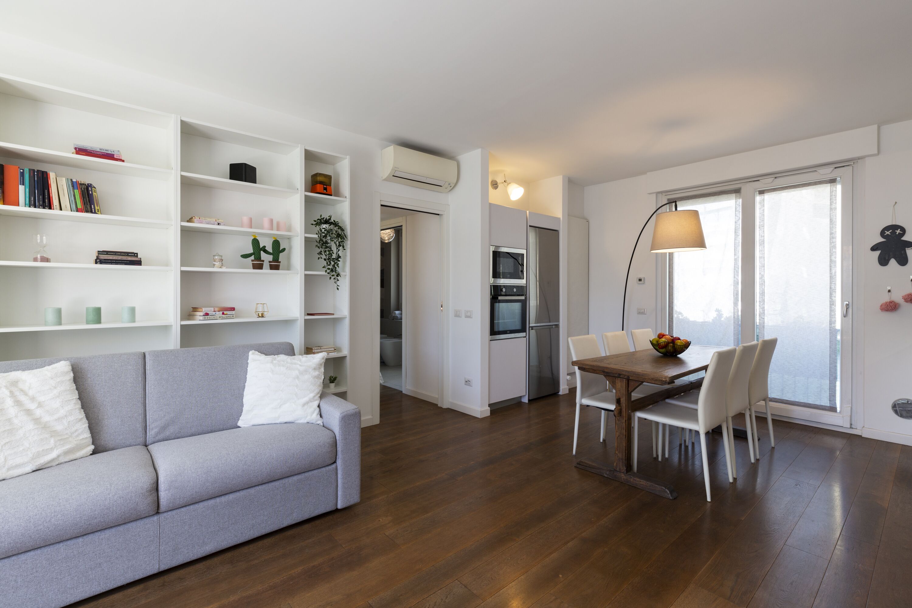 A modern living room with a grey sofa, white cushions, a wooden dining table with chairs, a kitchen area with appliances, and a large window with a view.