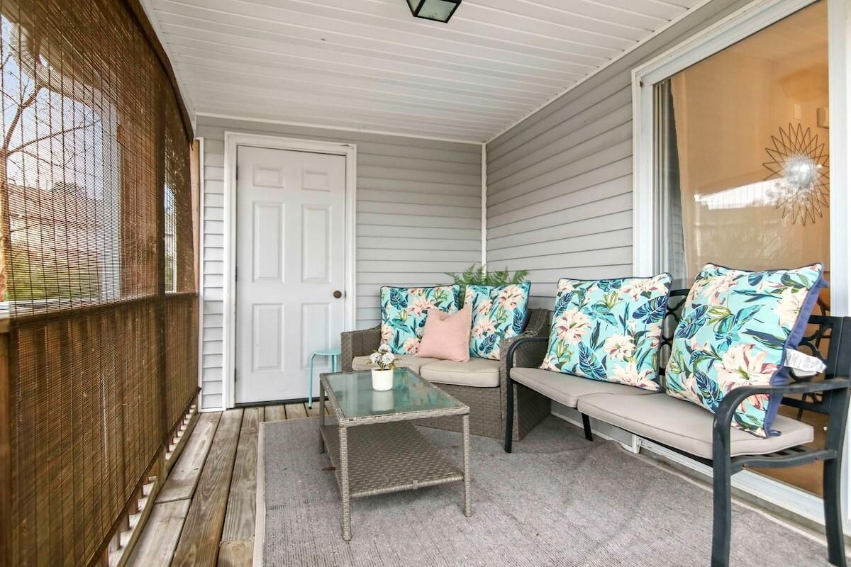 A covered porch with a wicker table, a cushioned bench with floral cushions, and a view of the outdoors.