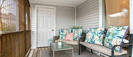 A covered porch with a wicker table, a cushioned bench with floral cushions, and a view of the outdoors.