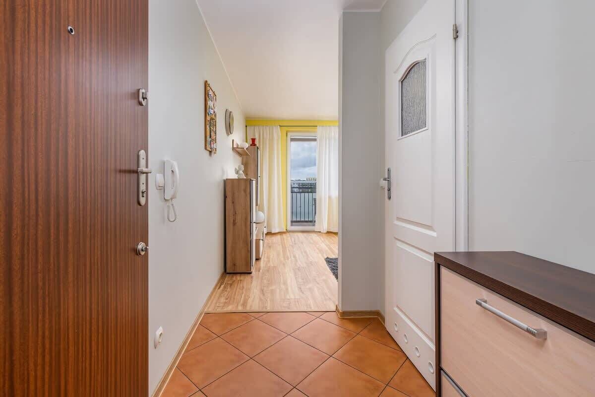 Ein Flur mit einer Holztür, einer weißen Wand, einem Holzschrank und Blick auf einen Balkon mit gelben Vorhängen.