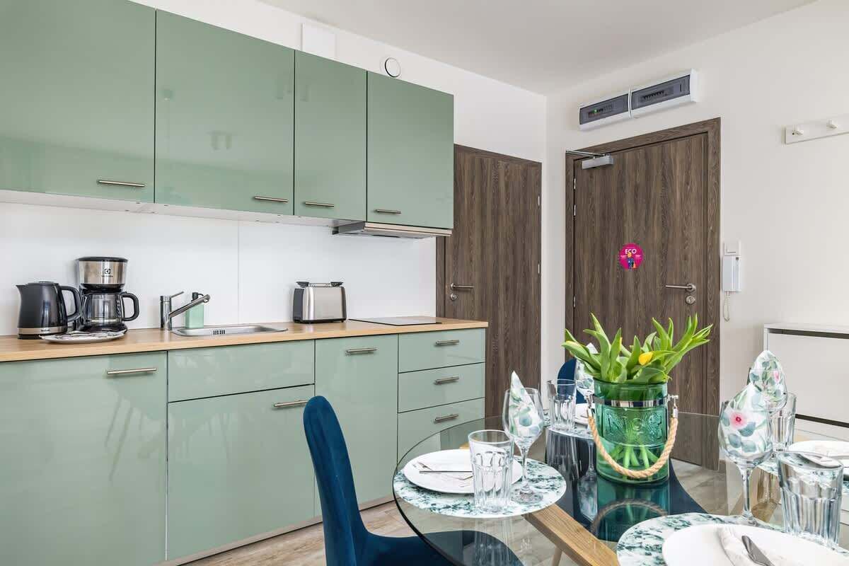 Une cuisine moderne avec des meubles de couleur verte, une machine à café et une table à manger agrémentée de beaux verres et d’une plante décorative.