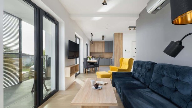 Uma sala de estar moderna com um sofá azul escuro, uma mesa de centro de madeira e uma poltrona amarela. Há uma cozinha ao fundo, com armários de madeira e uma mesa de jantar.