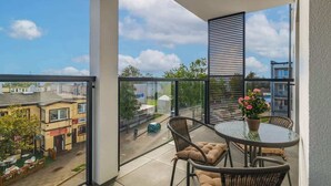 Uma varanda com mesa e cadeiras, um vaso de planta e vista para a rua e prédios.