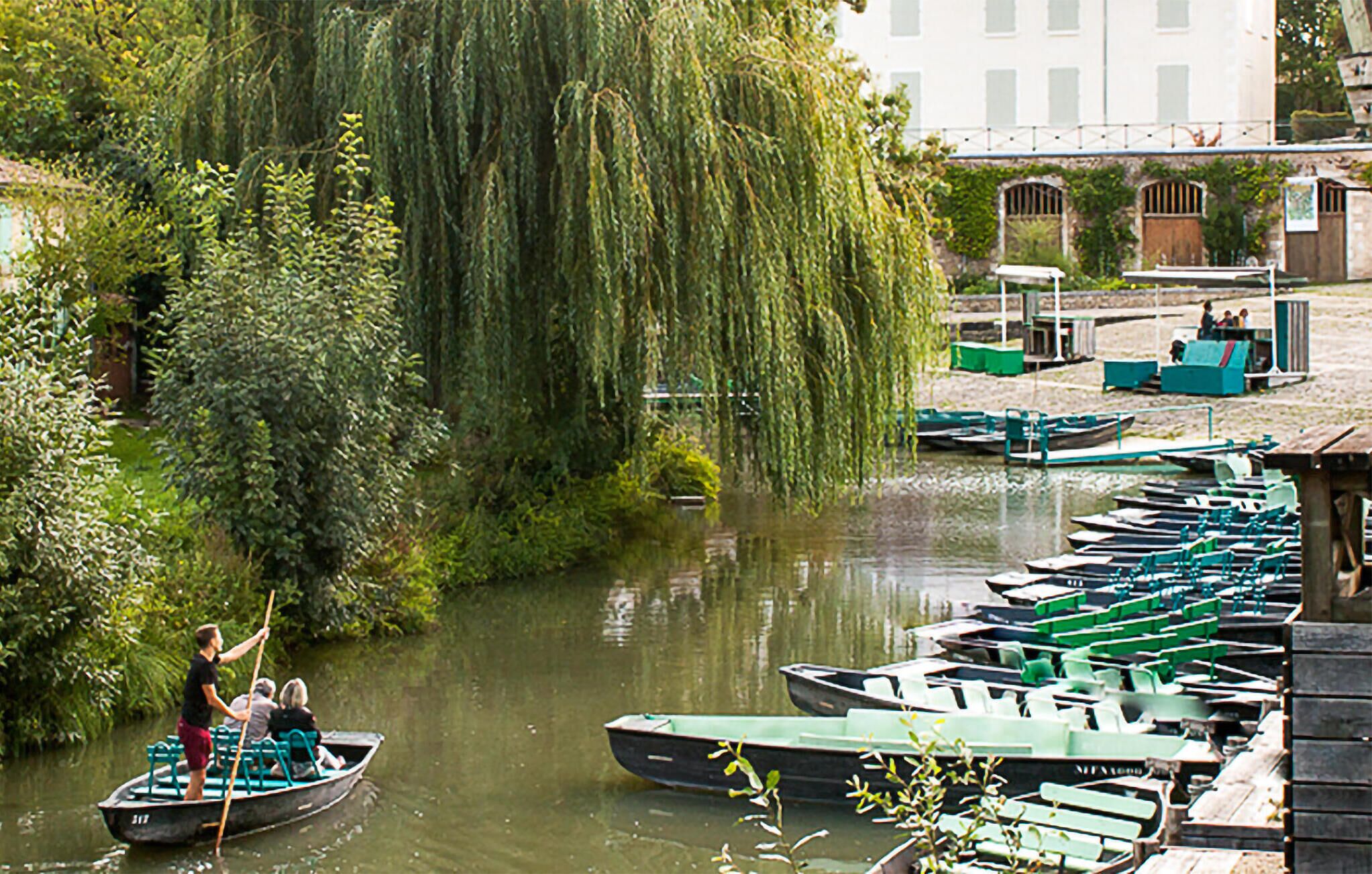 Ein Fluss mit vertäuten Booten am Ufer, eine Trauerweide und im Hintergrund ein Gebäude mit Rundbogenfenstern.