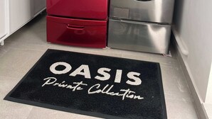 A laundry room with two front-loading washing machines, one red and one silver, and a black doormat with "OASIS Private Collection" written on it.
