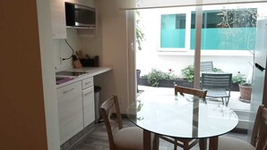 A compact kitchen with a microwave, sink, and cabinets. Outside, there's a glass door leading to a patio with a round table and chairs, and a view of a building with a green fence.