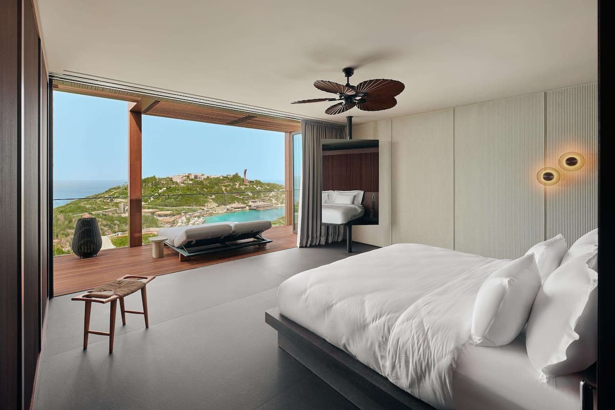 A modern bedroom with a large bed, a ceiling fan, a view of the sea, and a balcony with a chair.