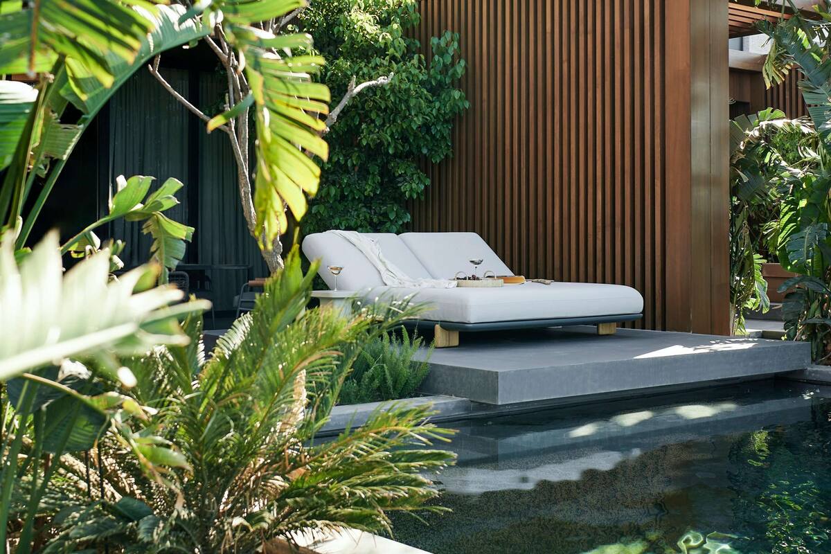 A modern outdoor lounge area with a white sofa, a wooden slatted wall, and lush tropical plants.