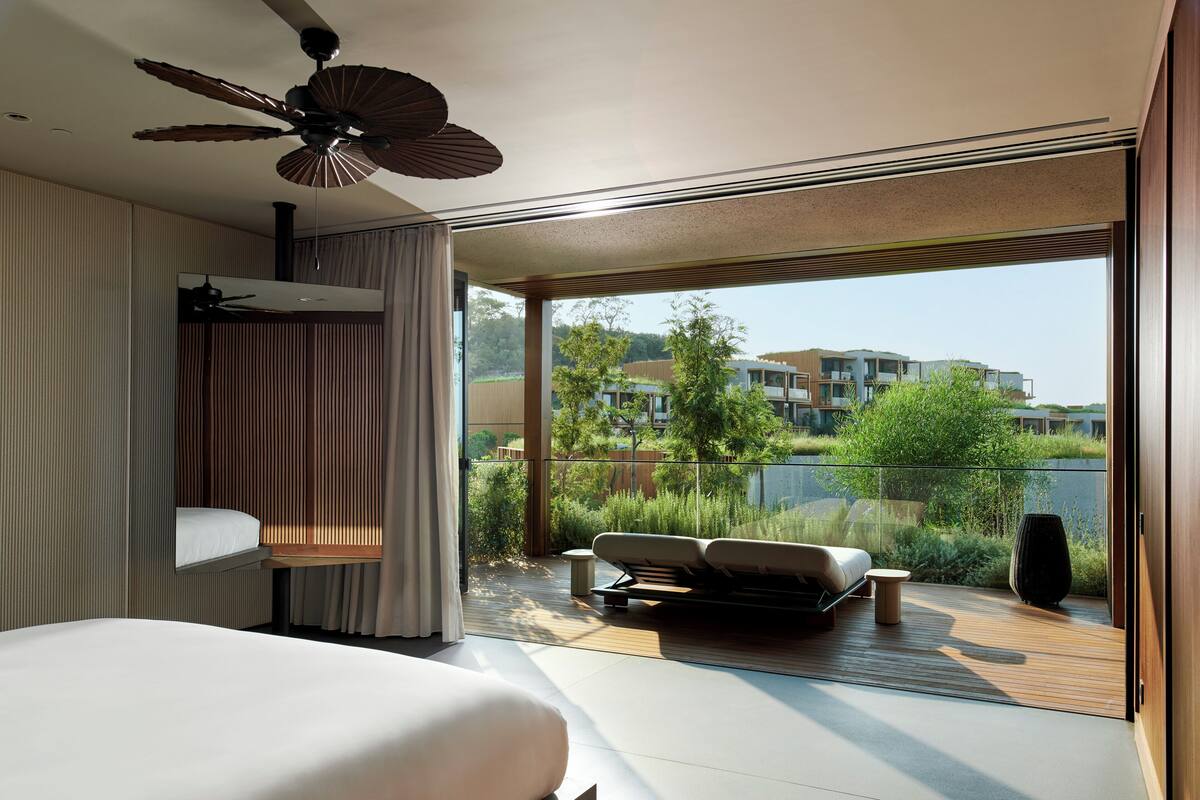 A modern bedroom with a large bed, a ceiling fan, and a view of a balcony with a sofa and a potted plant.