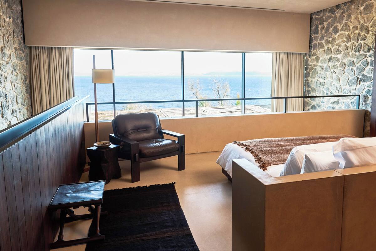 A hotel room with a bed, a chair, a small table, and a view of the sea through large windows.