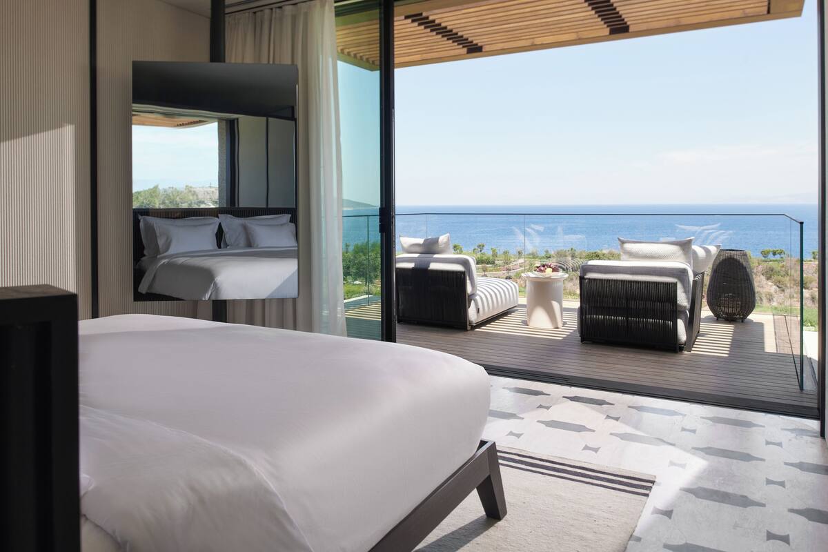A modern hotel room with a large bed, a balcony with seating, and a view of the sea.