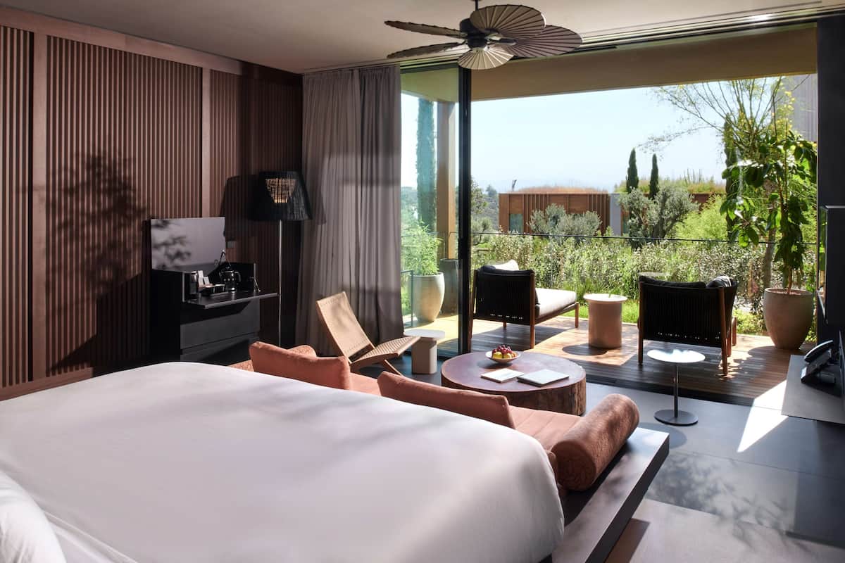 A modern hotel room with a large bed, a desk with a computer, a chair, a small table with fruit, and a balcony with plants and a view of the city.