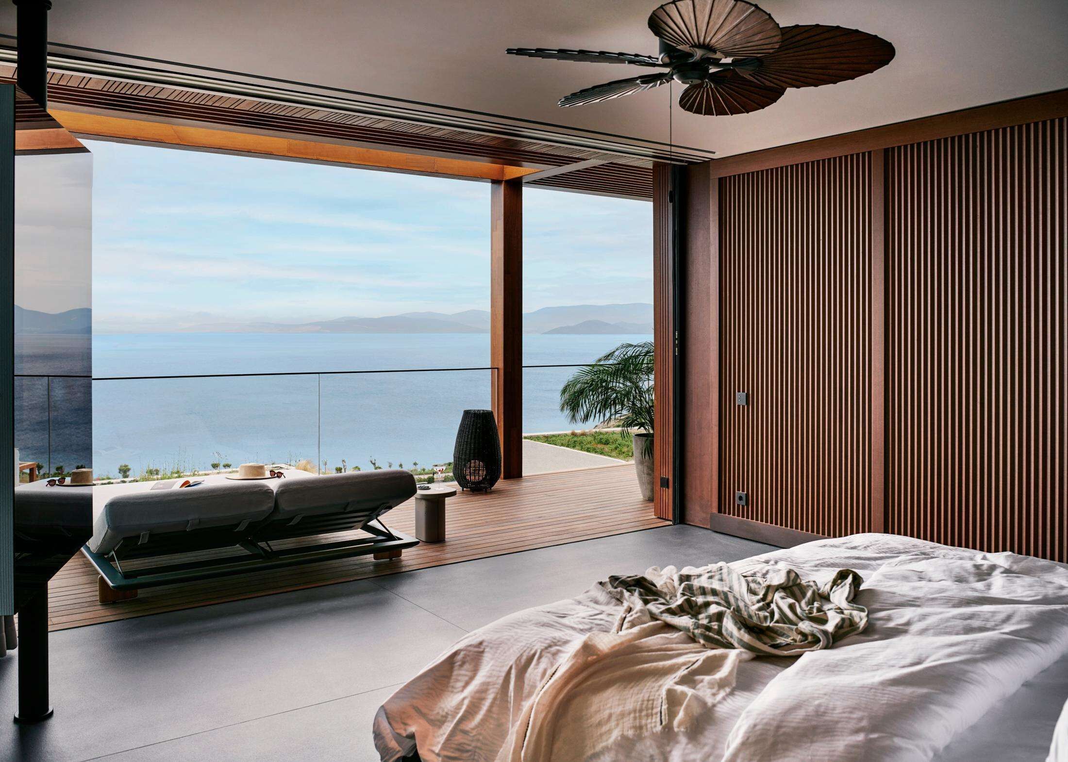 A modern bedroom with a large bed, a ceiling fan, and a view of the sea.