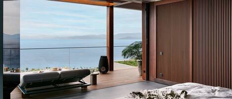 A modern bedroom with a large bed, a ceiling fan, and a view of the sea.