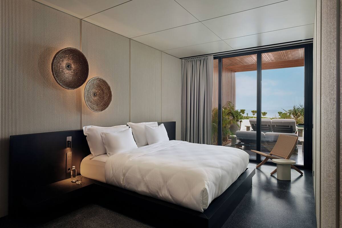 A modern bedroom with a large bed, a bedside table with a lamp, and a view of the outdoors through a sliding glass door.