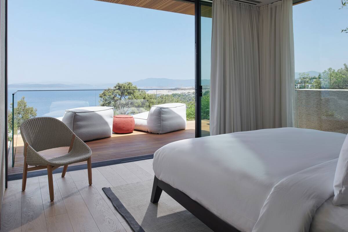 A modern hotel room with a large bed, a wicker chair, and a red ottoman. The room features a balcony with a view of the sea and mountains.
