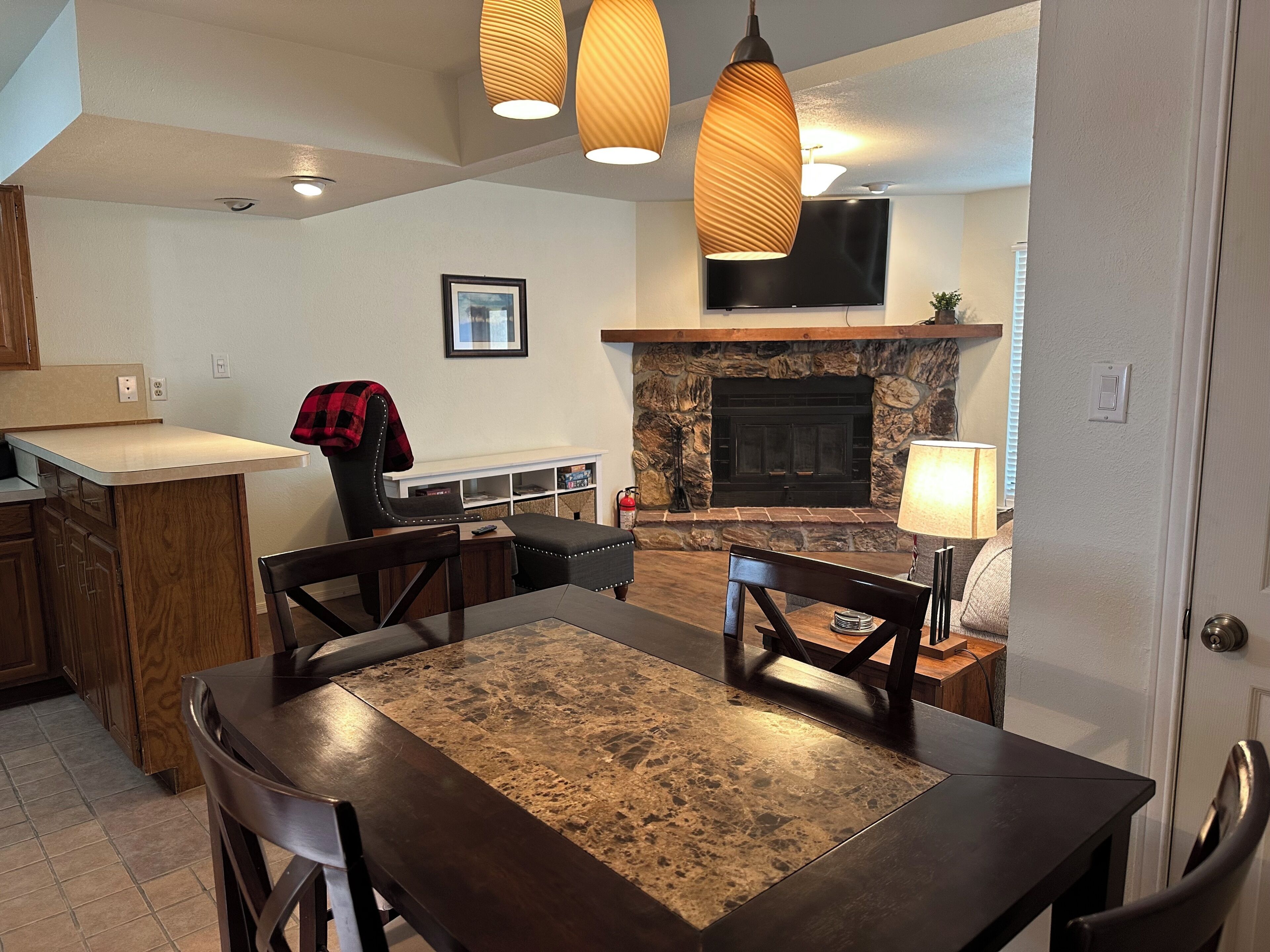 A dining area with a table and chairs, a kitchen with wooden cabinets, a stone fireplace, and pendant lighting.