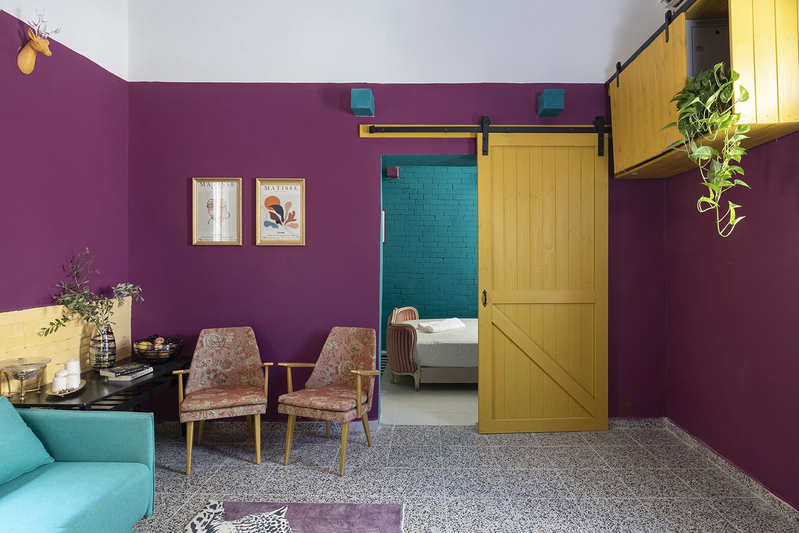 Quarto com paredes roxas, um sofá verde-água, duas cadeiras estampadas, uma cama com parede de tijolos azuis e uma porta de madeira.