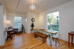 A spacious room with a wooden floor, a dining table with chairs, a bench, a lamp, a clock, and a large window.