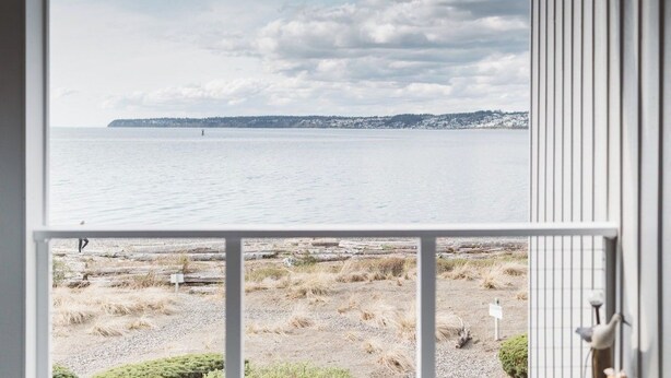 Une vue depuis une fenêtre donnant sur une étendue d’eau calme, avec une rive lointaine, une zone de gravier à la végétation clairsemée et une rambarde métallique sur la droite.