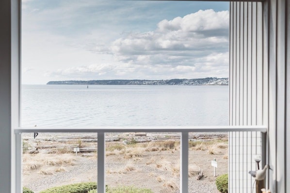 A view from a window overlooking a calm body of water with a distant shoreline, a gravel area with sparse vegetation, and a metal railing on the right.