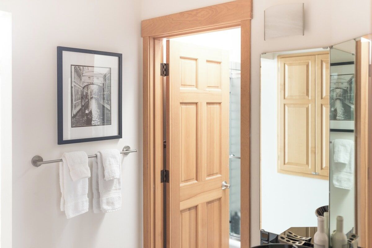 A bathroom with a wooden door, a towel rack with white towels, a framed picture on the wall, and a mirror reflecting a bathroom cabinet.