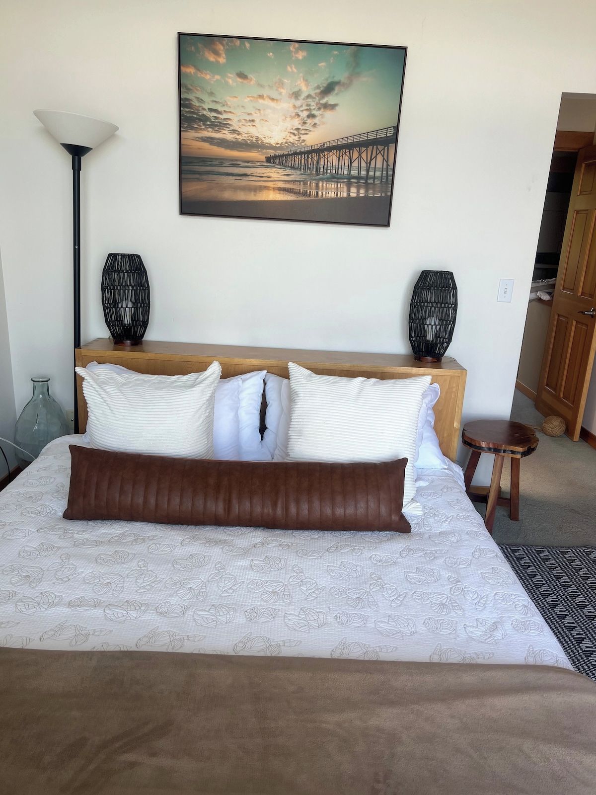 A bedroom with a bed, pillows, a nightstand, a lamp, a painting, and a door.