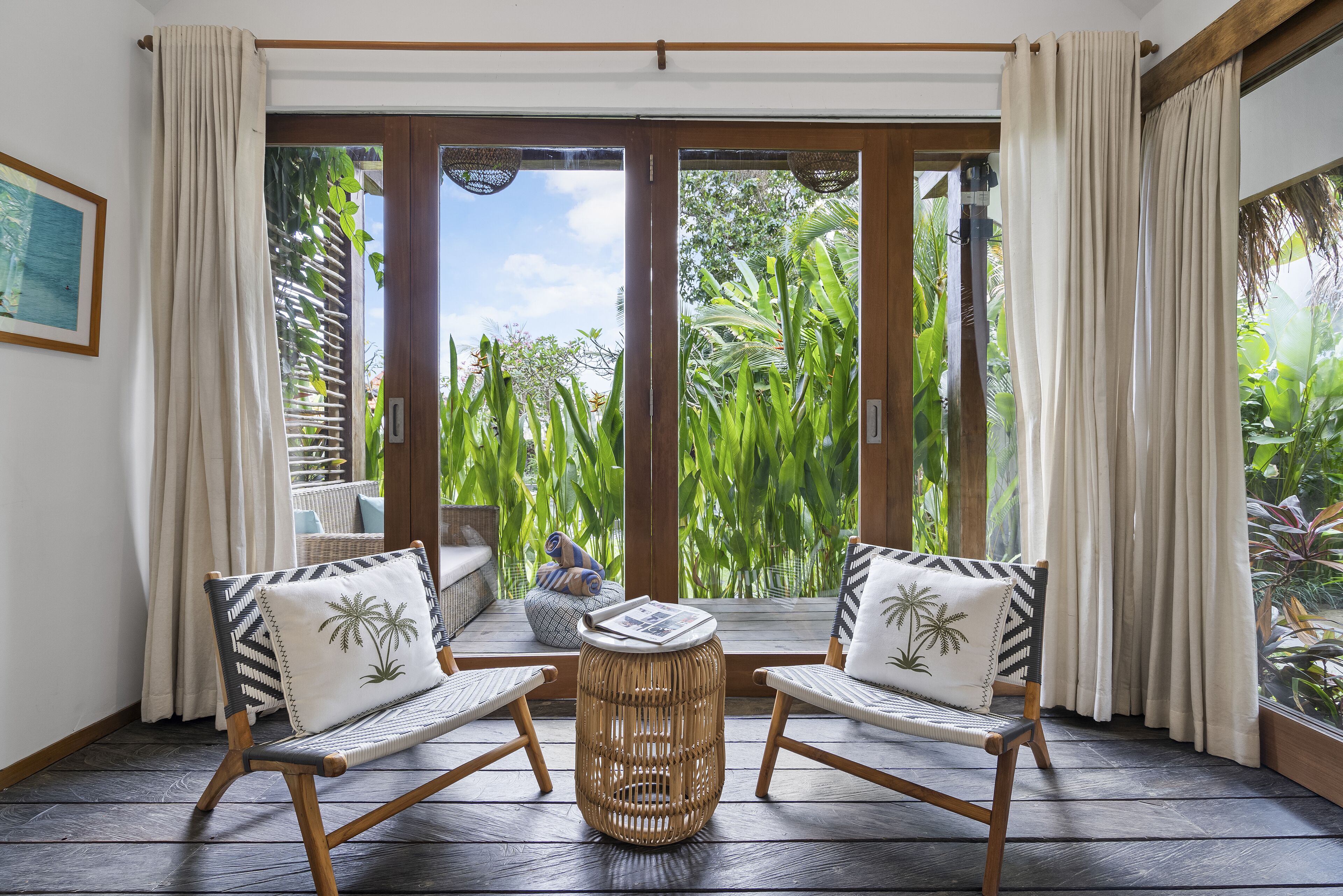 Une pièce avec deux fauteuils en osier, une petite table ronde et une vue sur une végétation luxuriante, visible à travers de grandes portes vitrées.