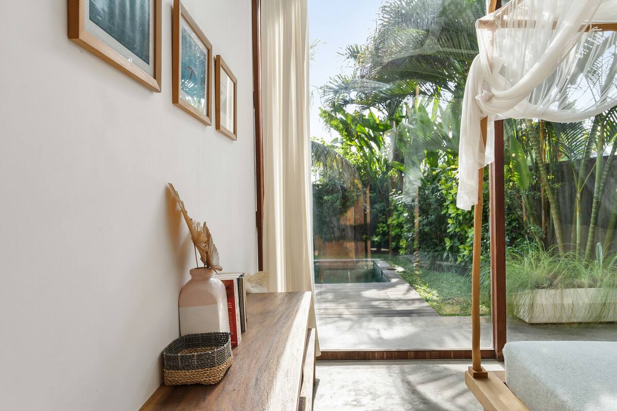 A room with a wooden bench, framed pictures, and a view of a garden through a glass door.