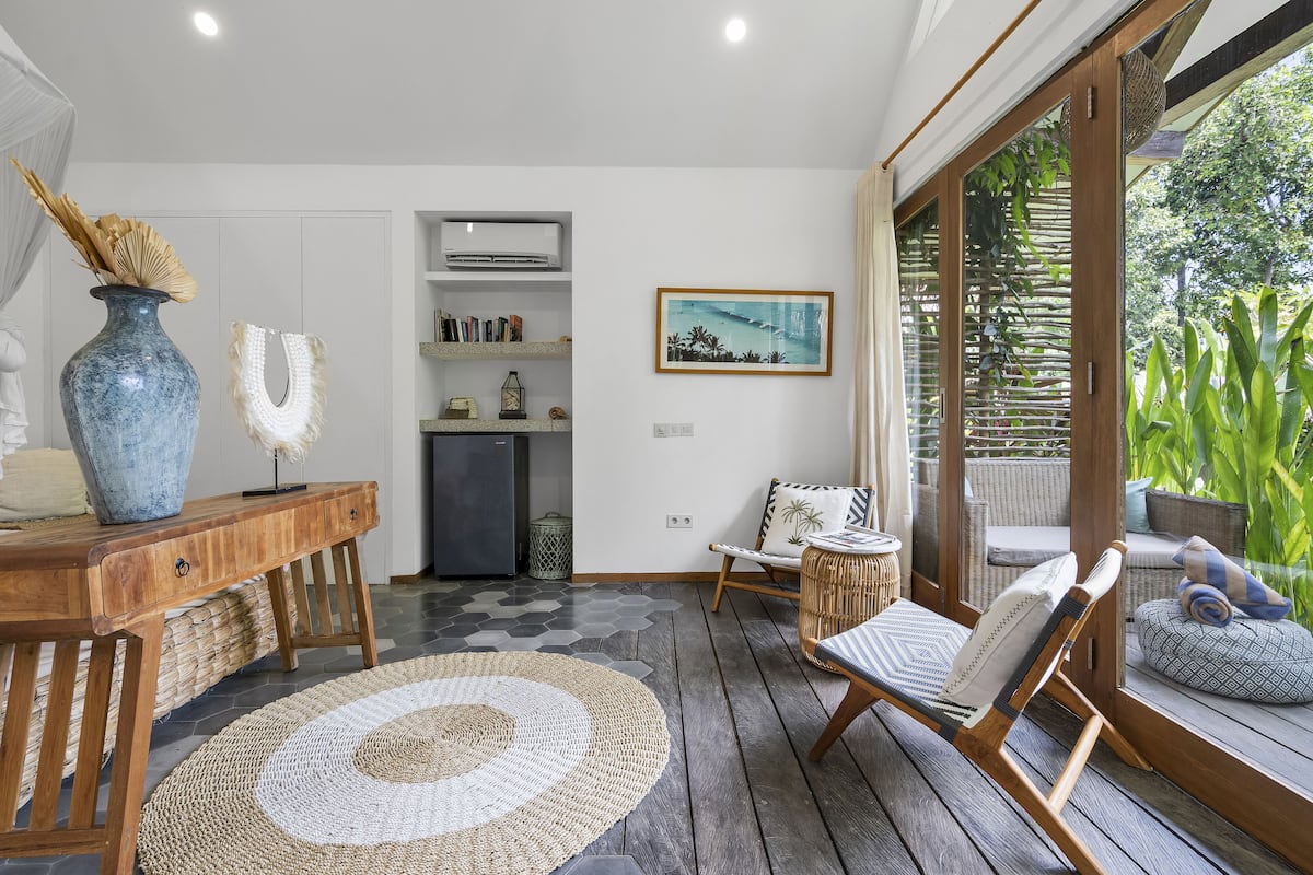 A cozy room with a wooden desk, a woven rug, a wicker chair, and a large window with a view of greenery.