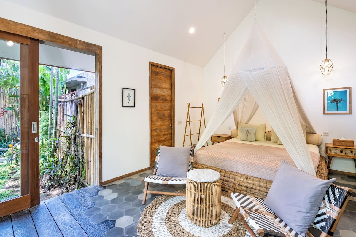 A bedroom with a canopy bed, a sitting area with a round table, and a view of a bamboo garden through a glass door.
