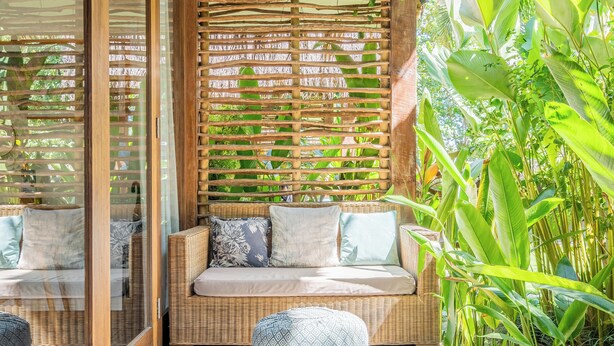 A wooden deck with a wicker sofa and ottoman, surrounded by lush greenery and a woven wall. - Ohana Bungalow by Betterplace  (Canggu)