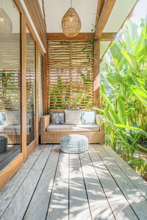 Una terraza de madera con un sofá y reposapiés de mimbre, rodeada de exuberante follaje y una pared trenzada.
