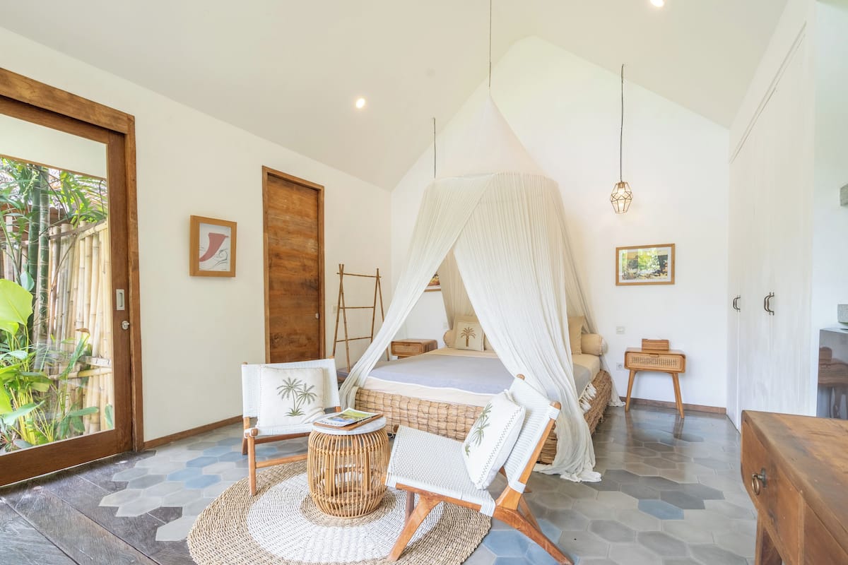 A bedroom with a canopy bed, a wicker chair, a small round table, and a view of greenery outside.