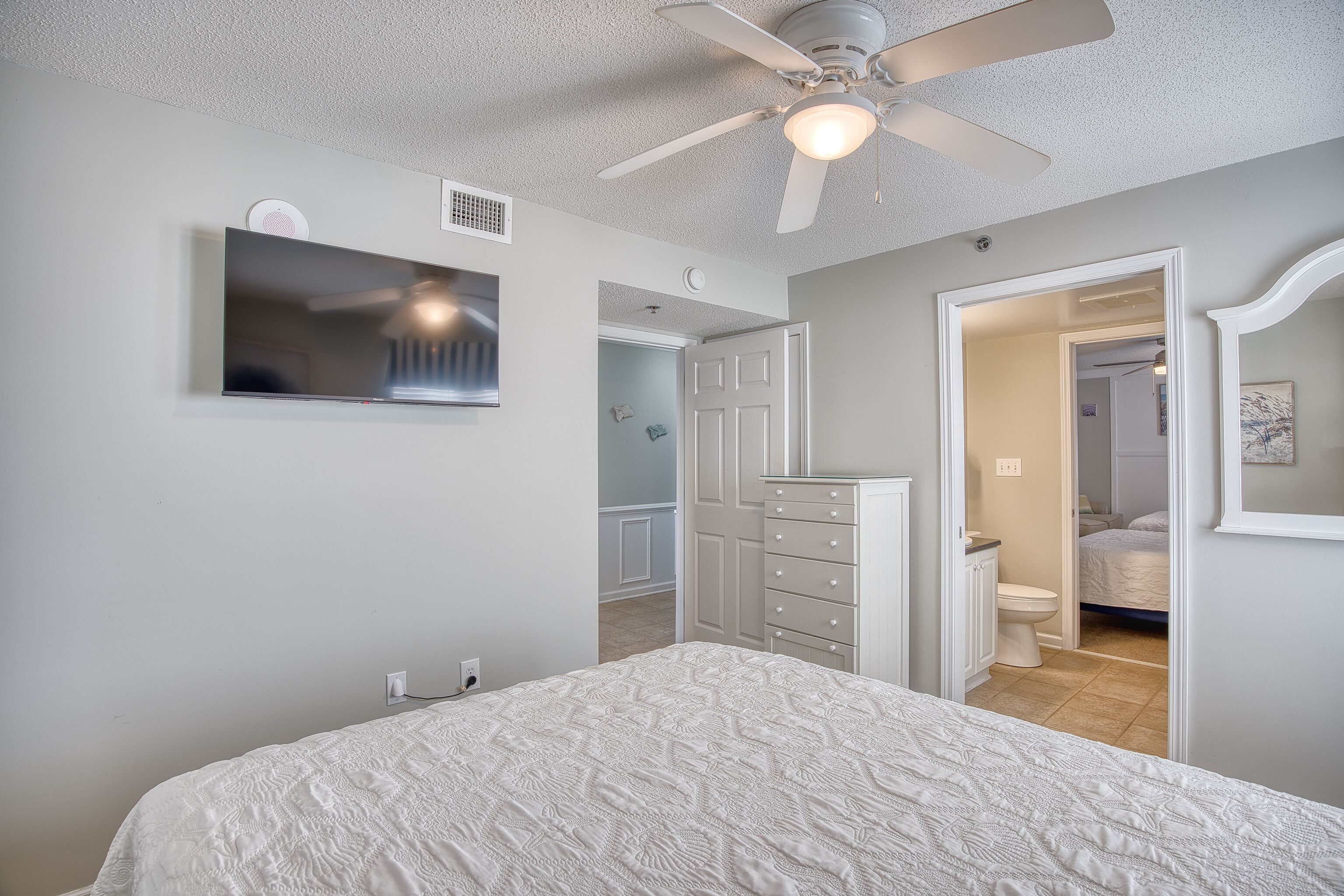 Une chambre à coucher comprenant un lit, un ventilateur de plafond, un téléviseur fixé au mur, une commode et une porte donnant accès à une salle de bain.