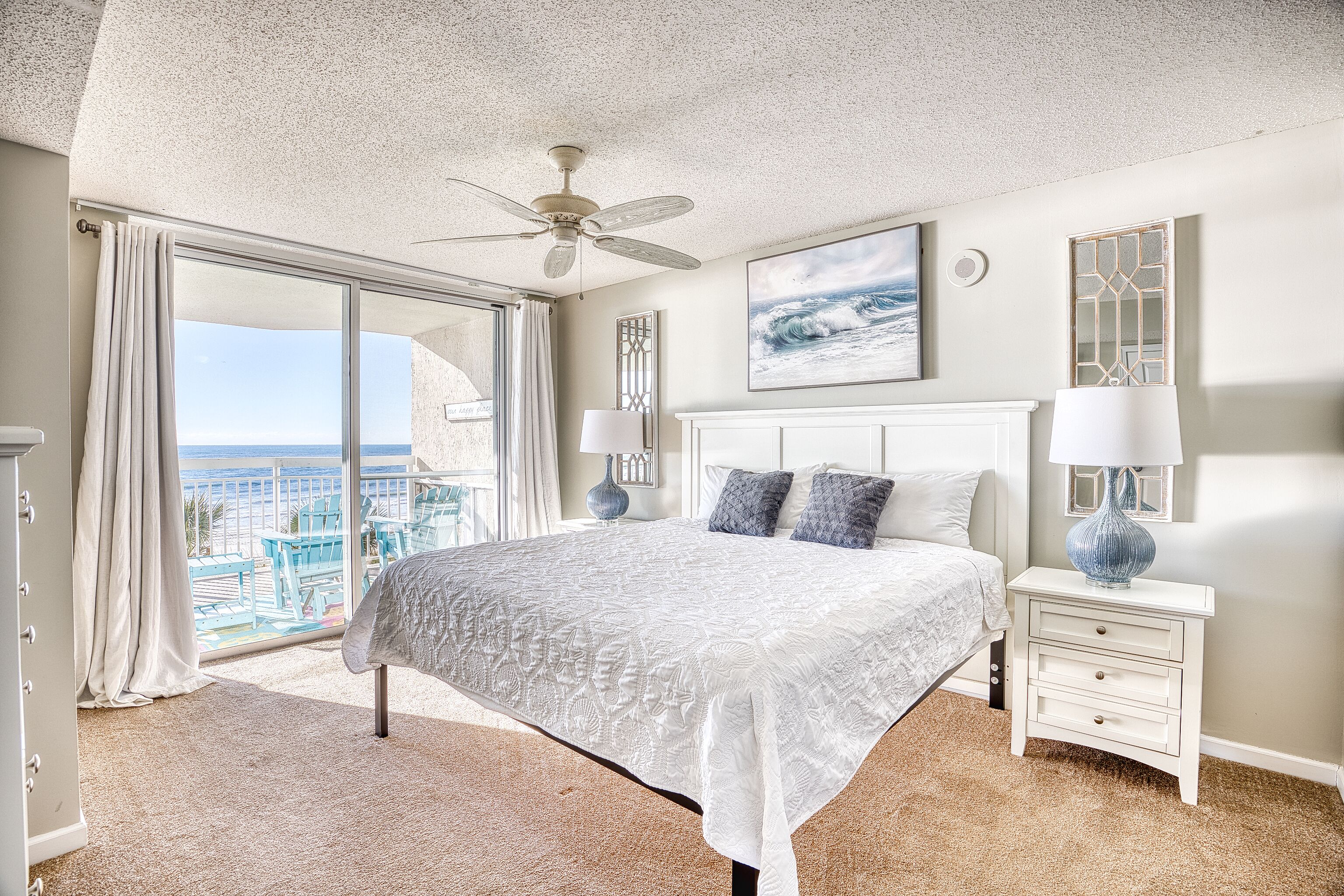 A bedroom with a large bed, a ceiling fan, a nightstand with a lamp, a view of the ocean through a sliding door, and a framed picture on the wall.