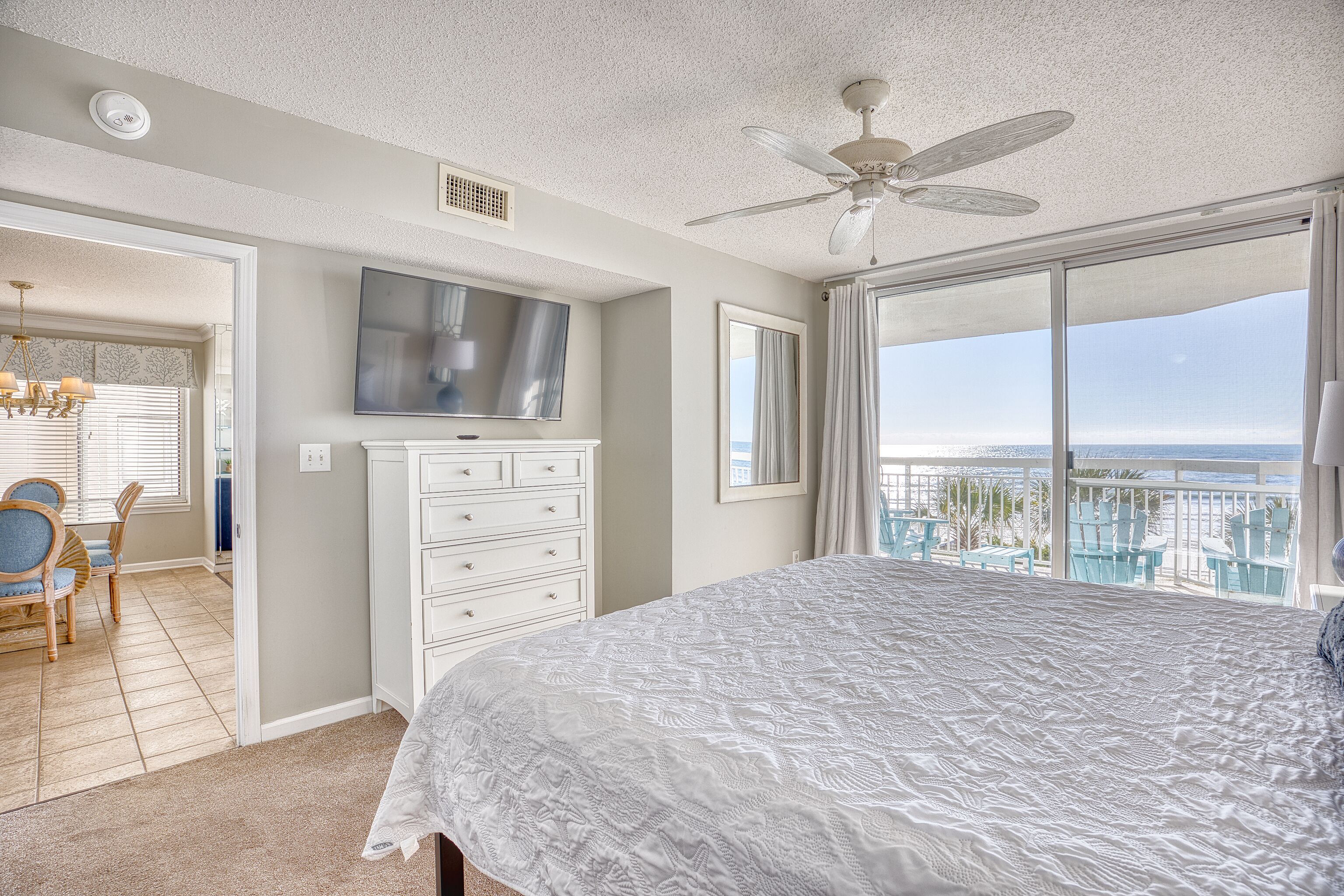 A bedroom with a large bed, a ceiling fan, a TV, and a balcony with a view of the ocean.