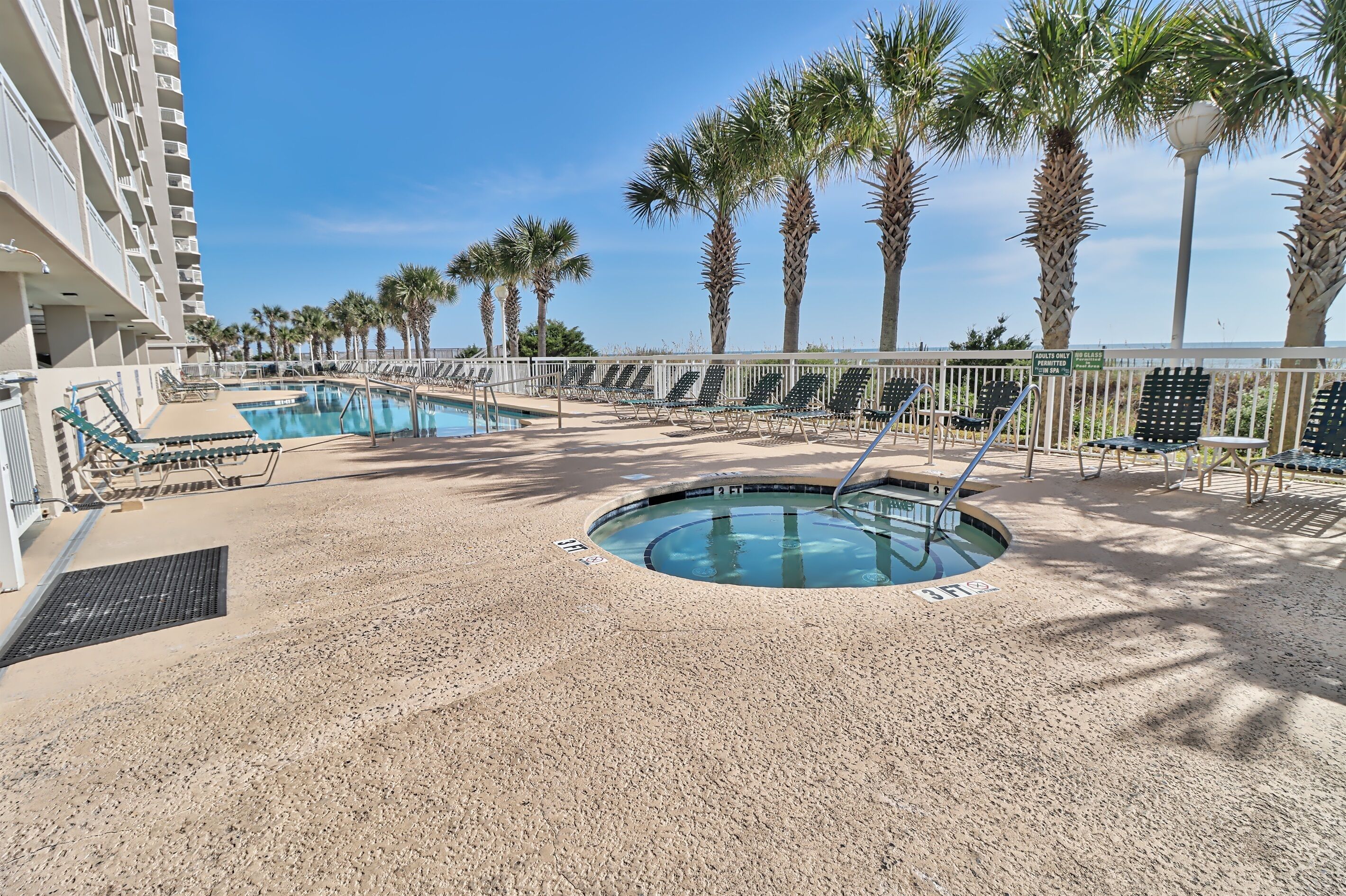 Zona de piscina con sillones reclinables, un jacuzzi y palmeras.