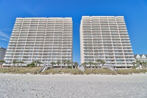 Two high-rise buildings with balconies, a sandy beach in the foreground, and palm trees. - Crescent Shores 206S | Oceanfront 4 Bedroom Condo (North Myrtle Beach)