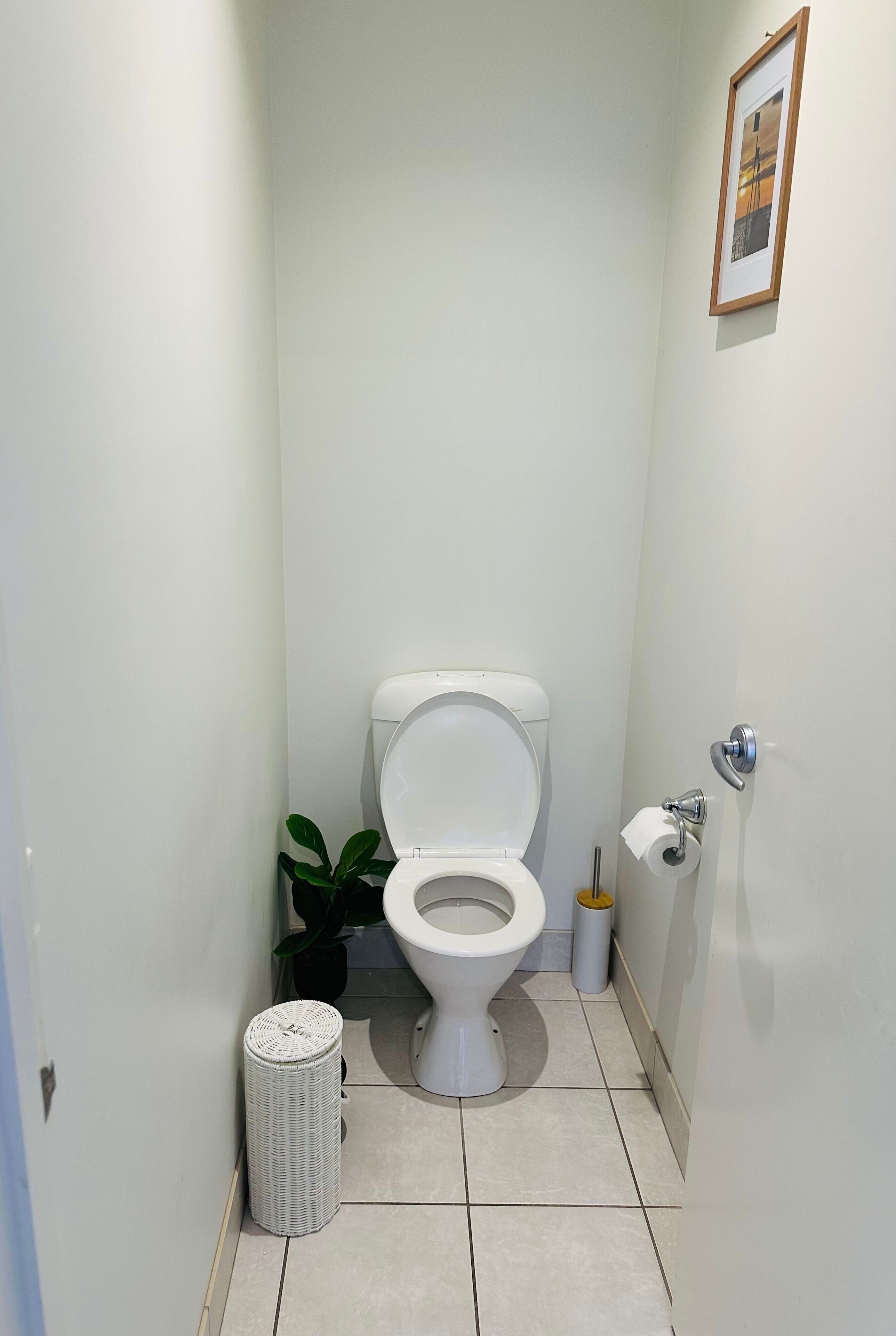 A small, clean bathroom with a toilet, a trash bin, a small potted plant, and a framed picture on the wall.