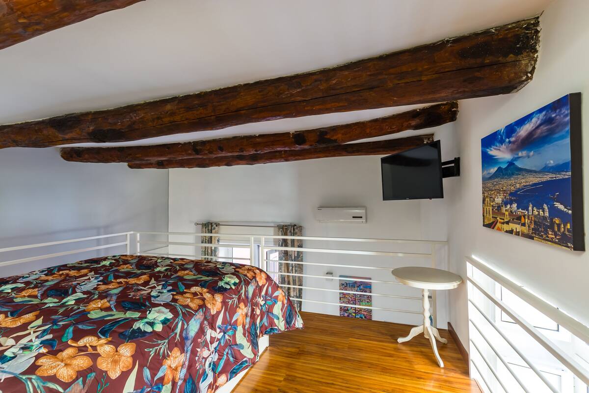 A bedroom with a wooden ceiling, a bed with a floral bedspread, a small table, a window with a cityscape view, and a mounted TV.